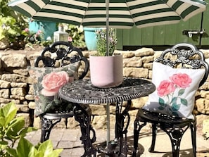 A round metal table with a potted plant, two chairs with floral cushions, and a striped umbrella. - Wind in the Willows Moreton in Marshes (Gloucestershire)