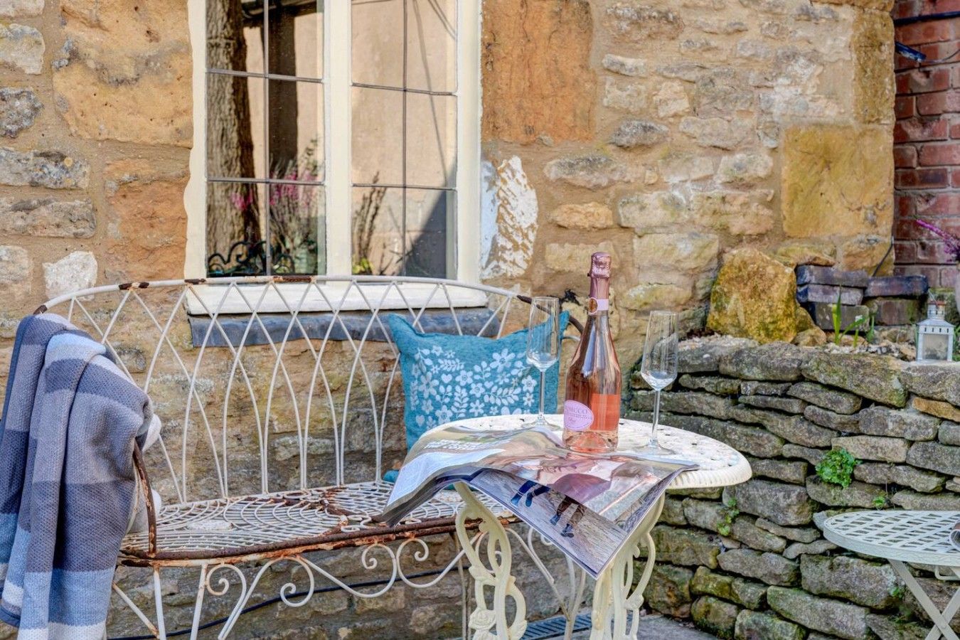 A rustic outdoor seating area with a metal chair, a table with a bottle and glasses, and a stone wall in the background.