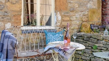 A rustic outdoor seating area with a metal chair, a table with a bottle and glasses, and a stone wall in the background.