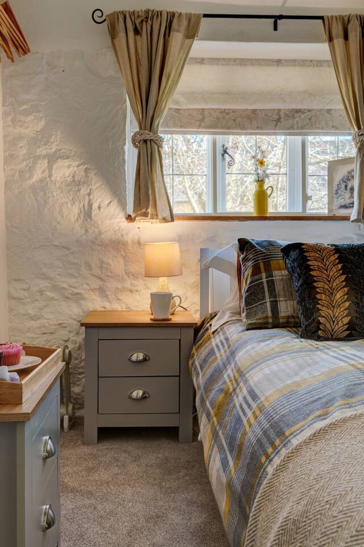 A bedroom with a bed, a nightstand with a lamp, a window with curtains, and a view of trees outside.