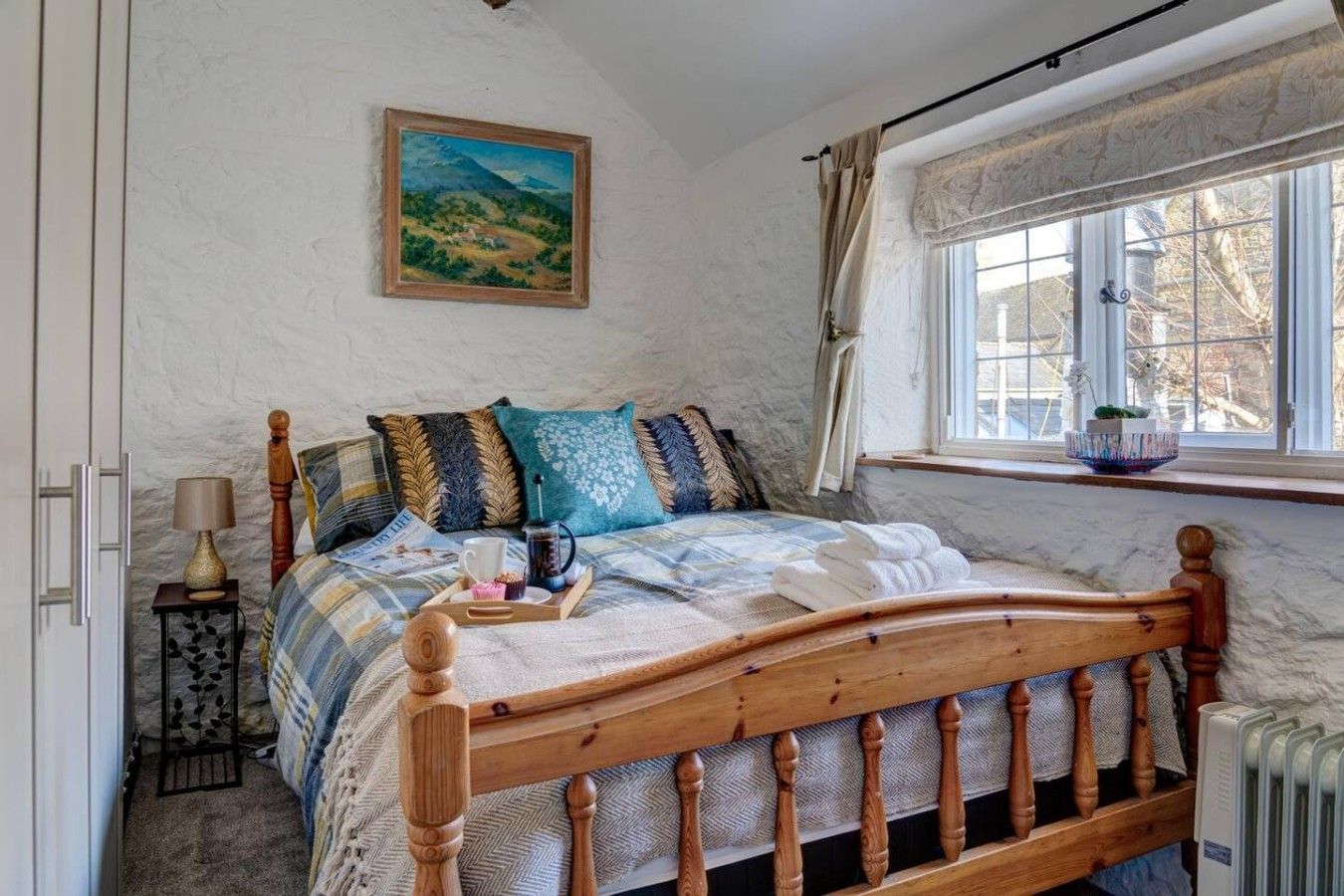 A bedroom with a wooden bed, a bedside table with a lamp, a window with curtains, and a painting on the wall.