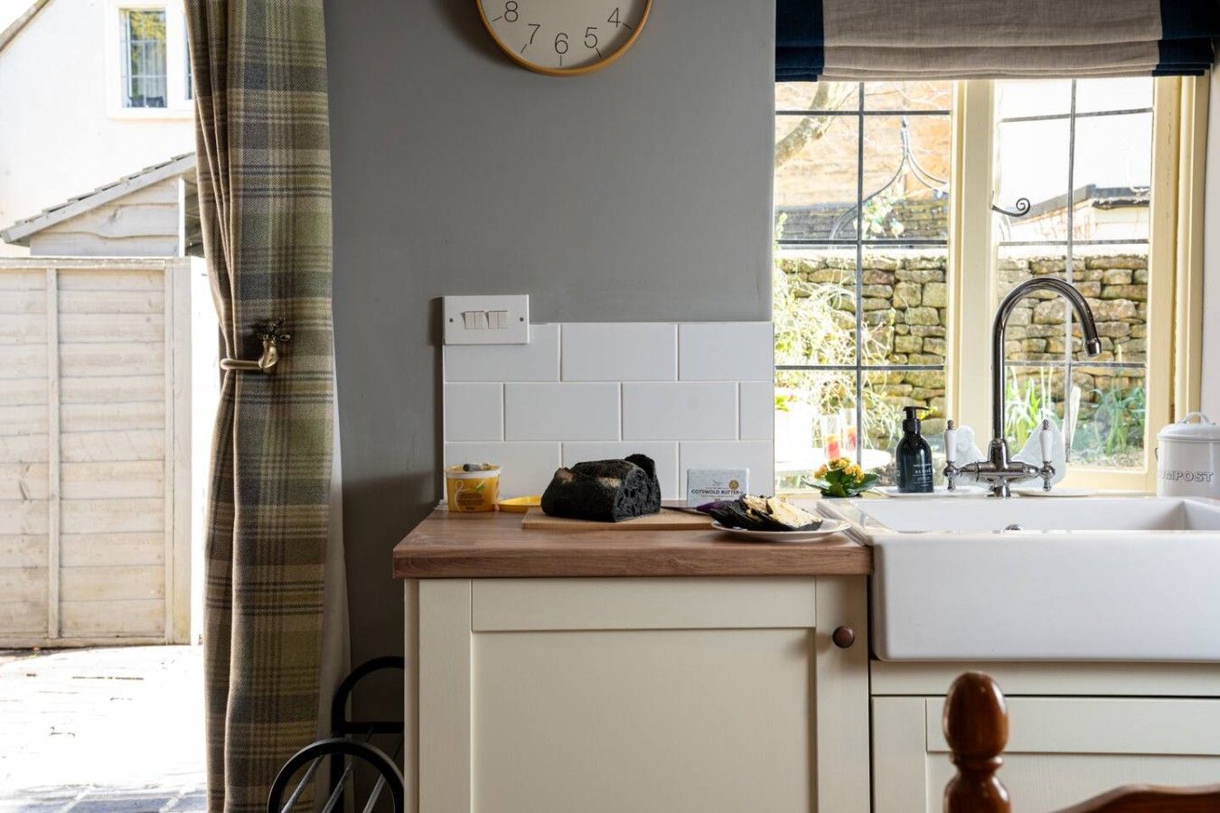 A kitchen with a wooden countertop, a clock on the wall, and a window with a view of a stone wall and greenery.
