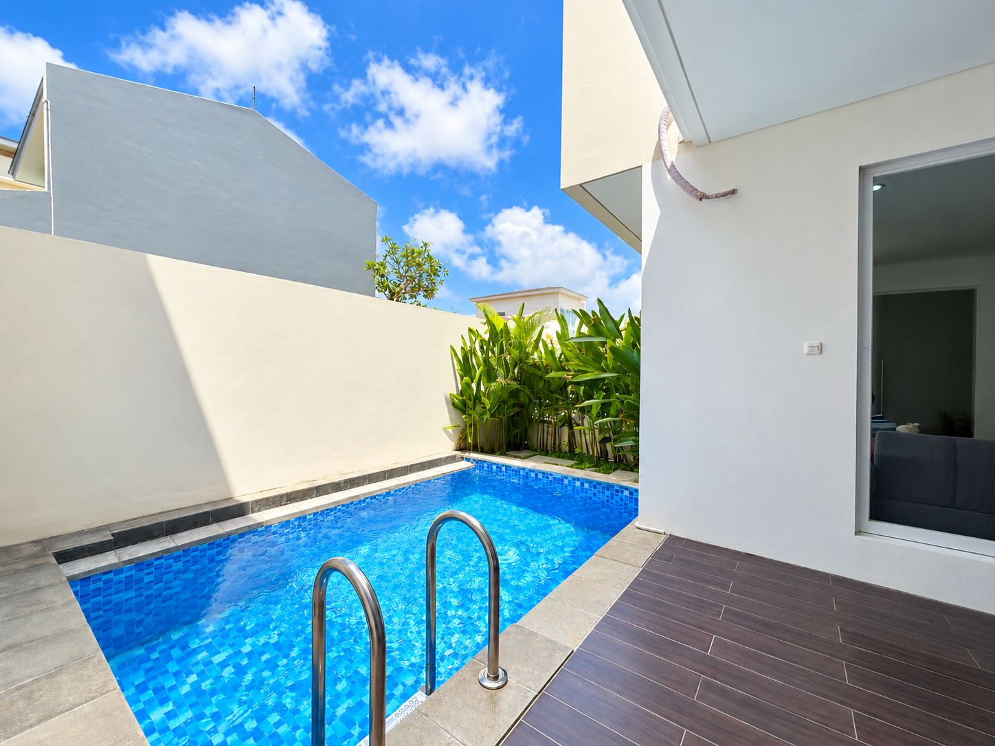 A modern outdoor swimming pool with a clear blue water surface, surrounded by a white wall and a dark wooden deck.
