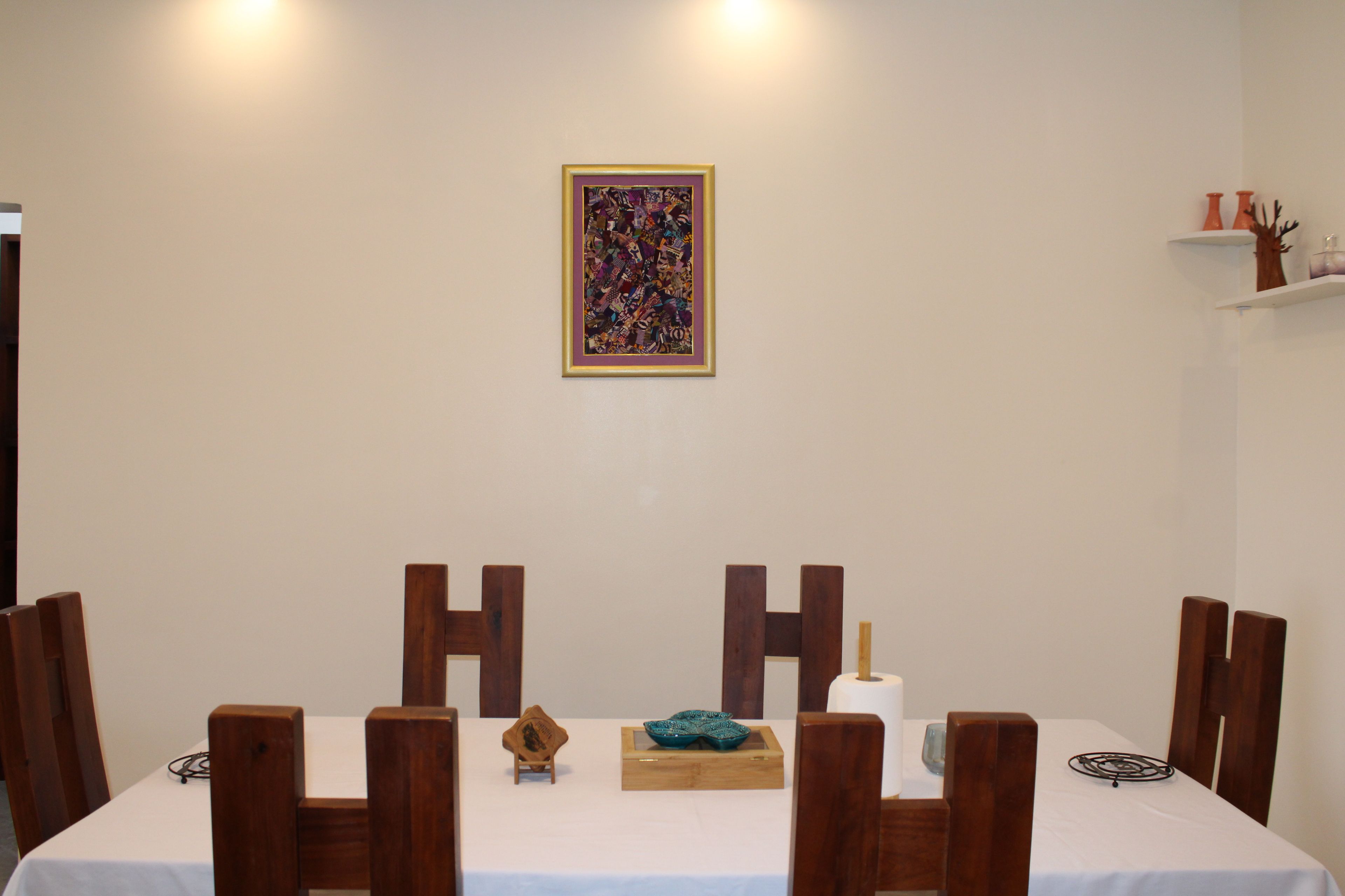 A dining area with a table set for four, wooden chairs, a framed abstract painting on the wall, and a small decorative item on the table.
