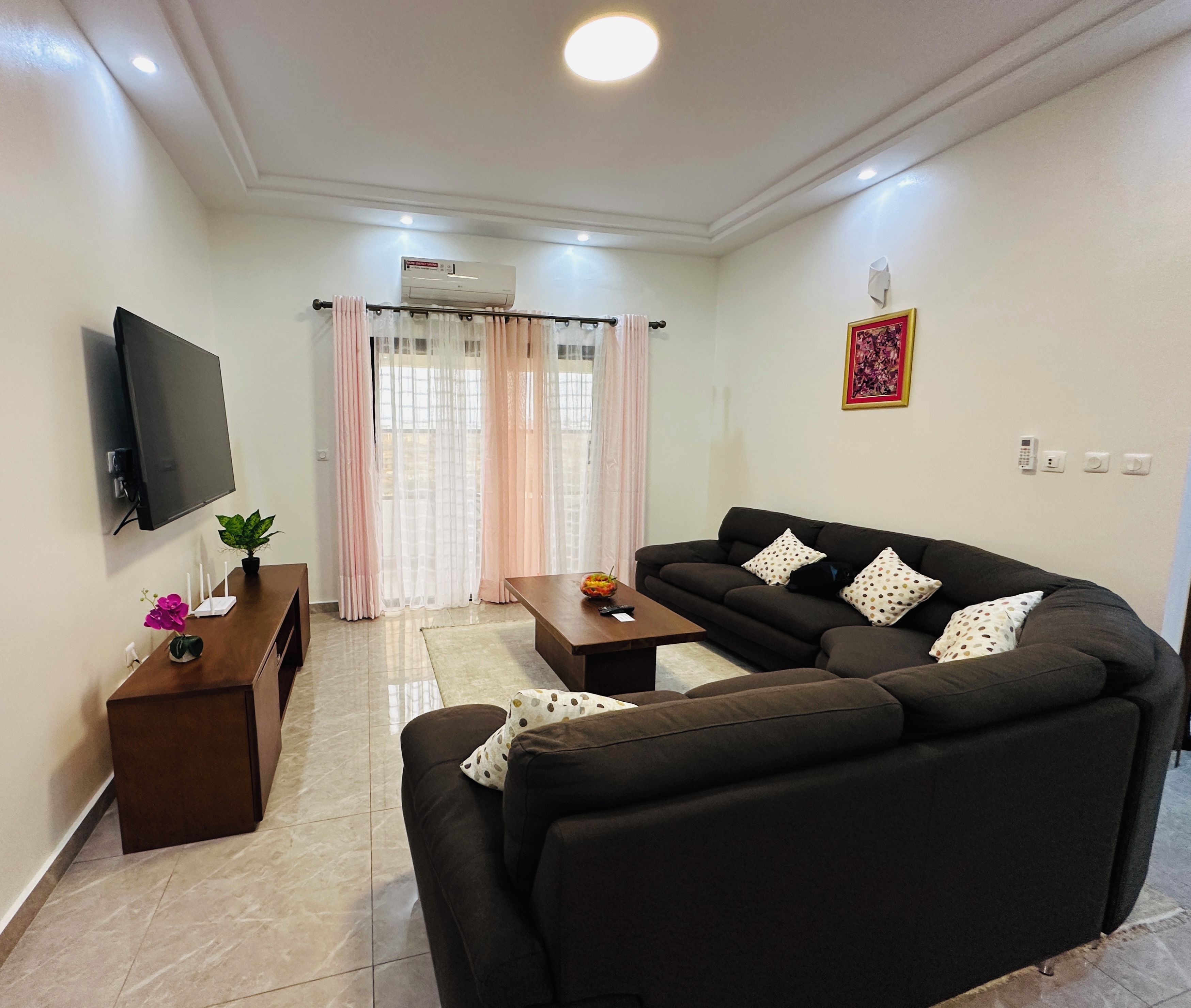 A living room with a sofa, a coffee table, a TV mounted on the wall, and a window with curtains.