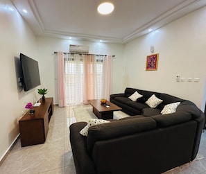 A living room with a sofa, a coffee table, a TV mounted on the wall, and a window with curtains. - Residence Nehemie (Abidjan)