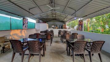 An outdoor dining area with tables and chairs, a ceiling fan, and a view of greenery.
