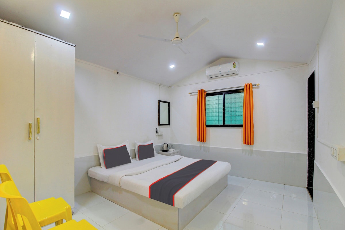 A hotel room with a bed, a wardrobe, a ceiling fan, an air conditioning unit, a window with curtains, and a small table with a lamp.