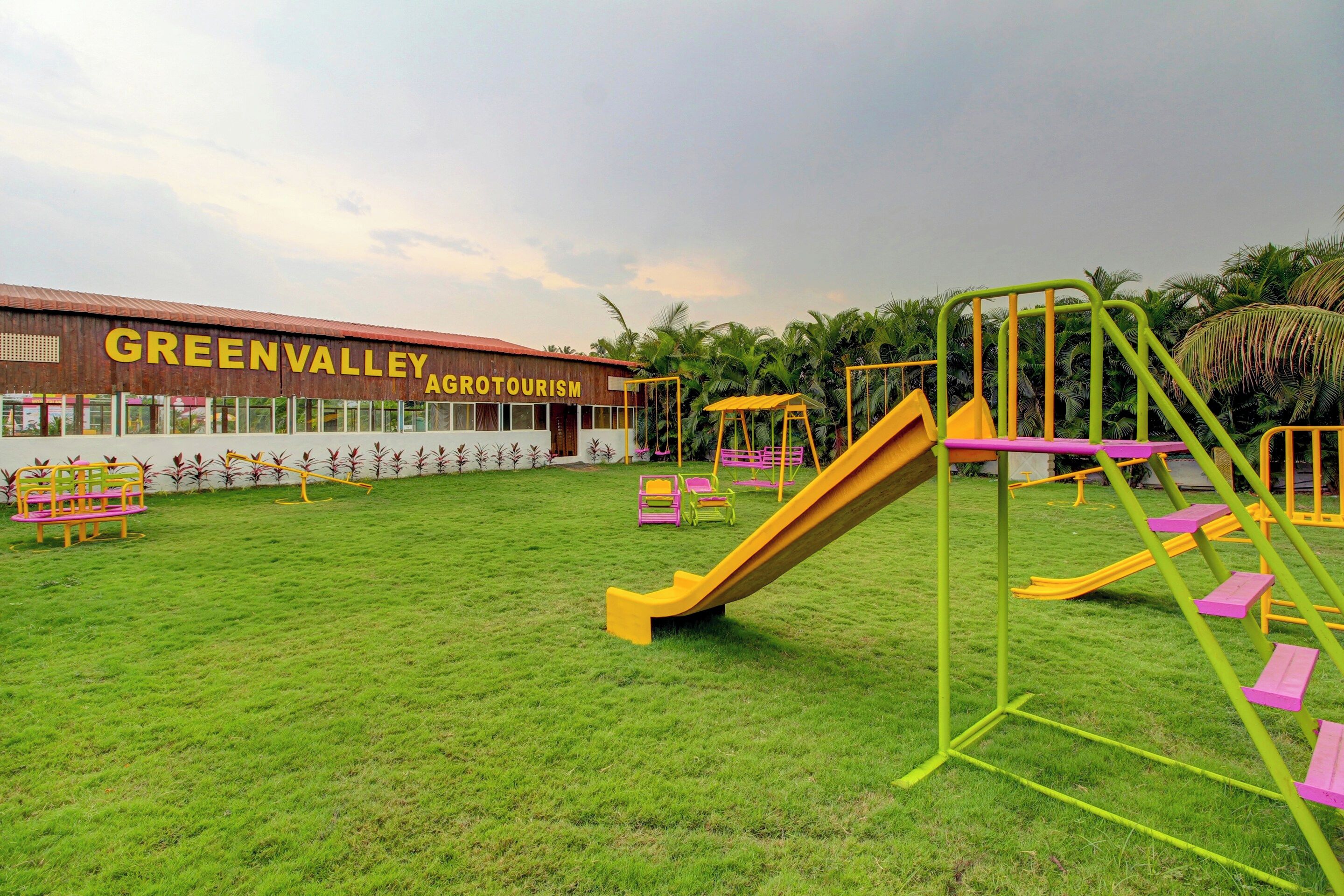 Ein Spielplatz mit Rutschen und Schaukeln, ein Gebäude mit der Beschriftung 'GREEN VALLEY AGRICULTURISM' und eine Rasenfläche.