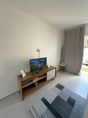 A modern living room with a flat-screen TV on a wooden stand, a small table with a vase, and a large rug on the floor. - New Listing (Cabo Frio)