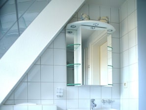 A bathroom with a white tiled wall, a white sink, a mirror with a curved frame, and a glass shelf. - Haus Bornholm Holiday Apartment Anemone (Nieblum)