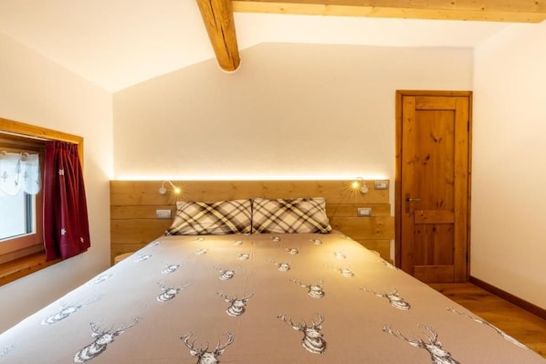 A bedroom with a bed, wooden headboard, a window with curtains, a door, and wall-mounted lights.