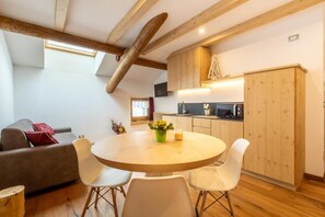 A modern kitchen with wooden cabinets, a dining table with chairs, and a sofa in a living area with a sloped ceiling. - Baita la Cascata  (Isolaccia)