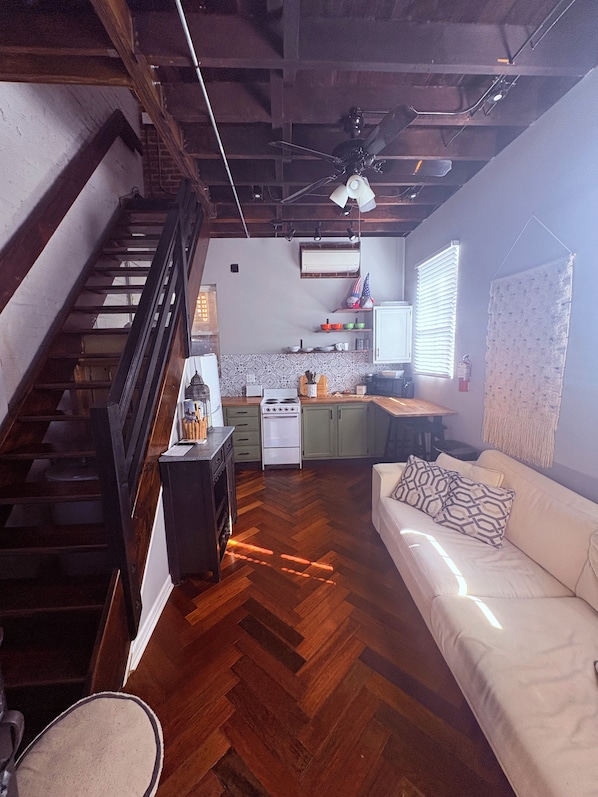 A spacious living area with a herringbone wood floor, a white sofa with patterned cushions, a kitchen with green cabinets, and a staircase with wooden railings.