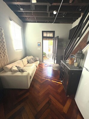A living room with a sofa, wooden flooring, a kitchen area with cabinets, and a door leading outside.