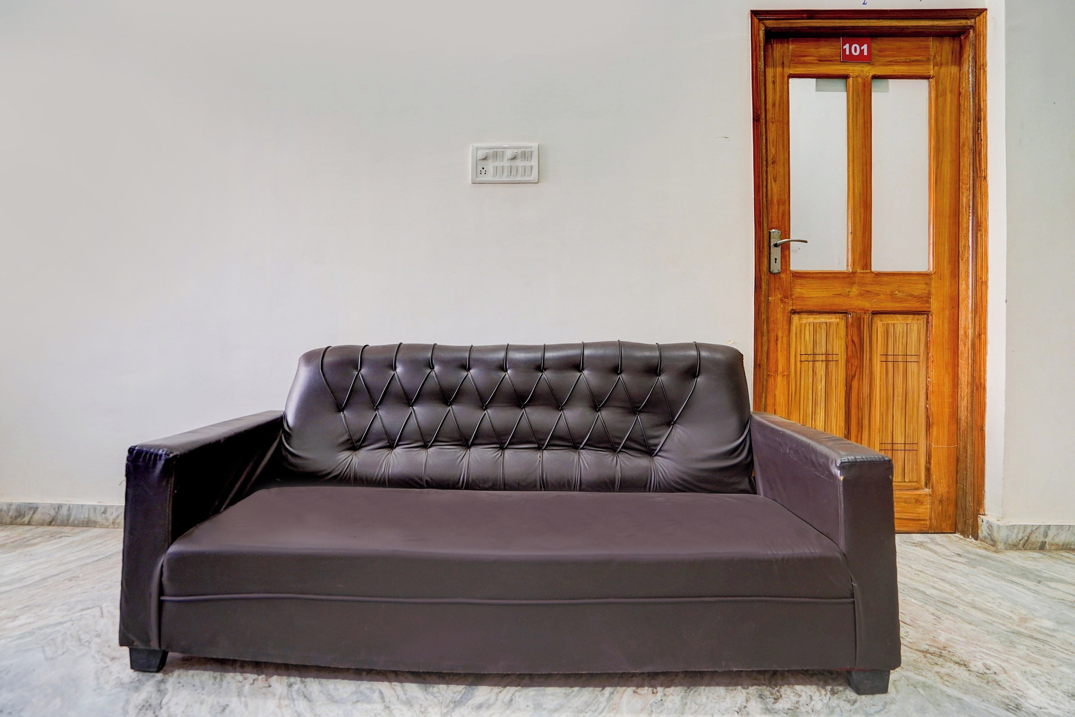 A dark brown leather sofa with a tufted backrest in a room with a wooden door and a marble floor.