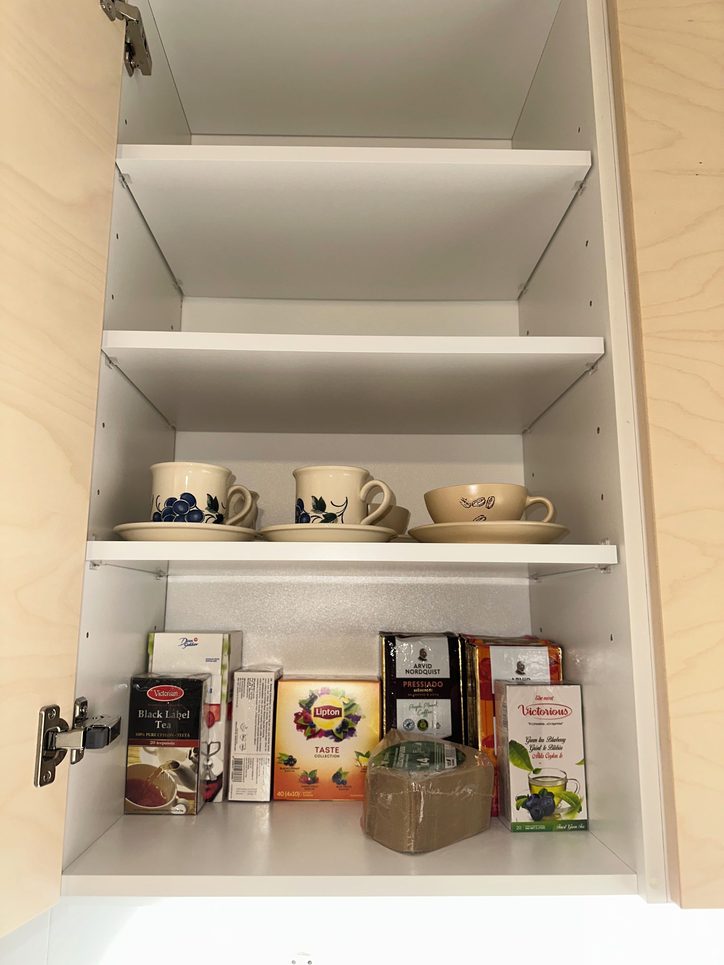Un meuble de cuisine doté d’étagères où sont rangées des boîtes de thé, une machine à café et un pot à sucre.