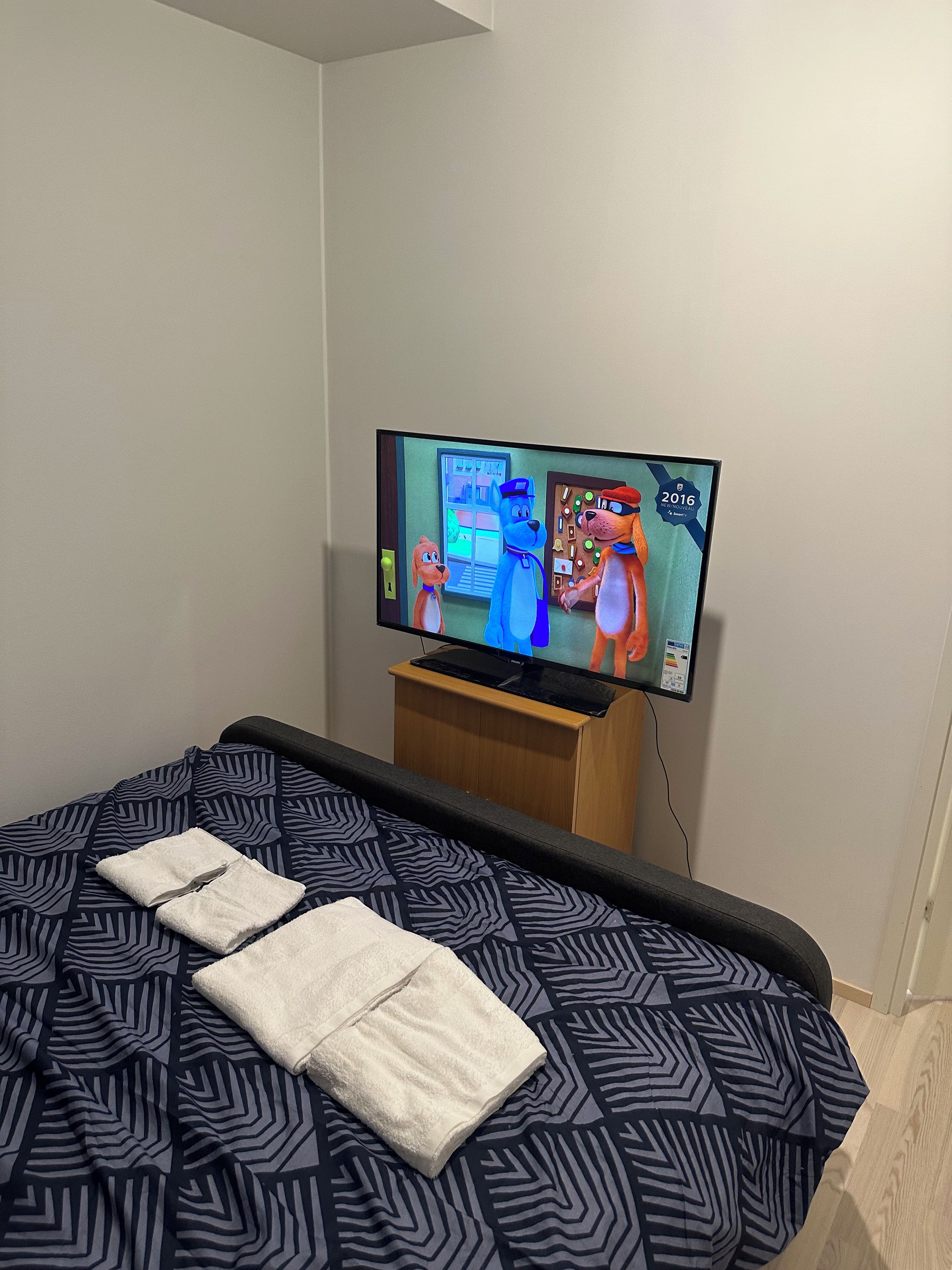 A hotel room with a bed, a television displaying animated characters, and a wooden nightstand.