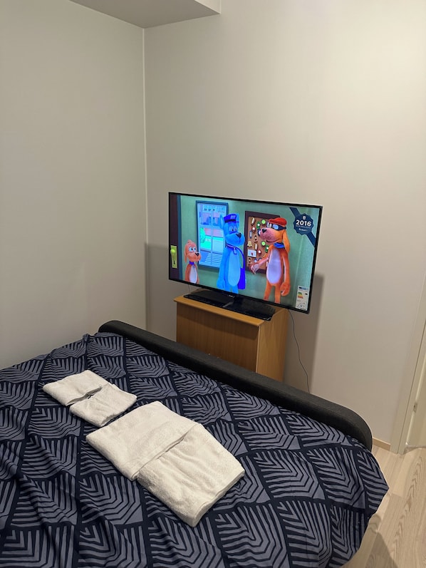 Hotellihuone, jossa on sänky, televisiossa näkyy piirrettyjä hahmoja ja huoneessa on puinen yöpöytä.
