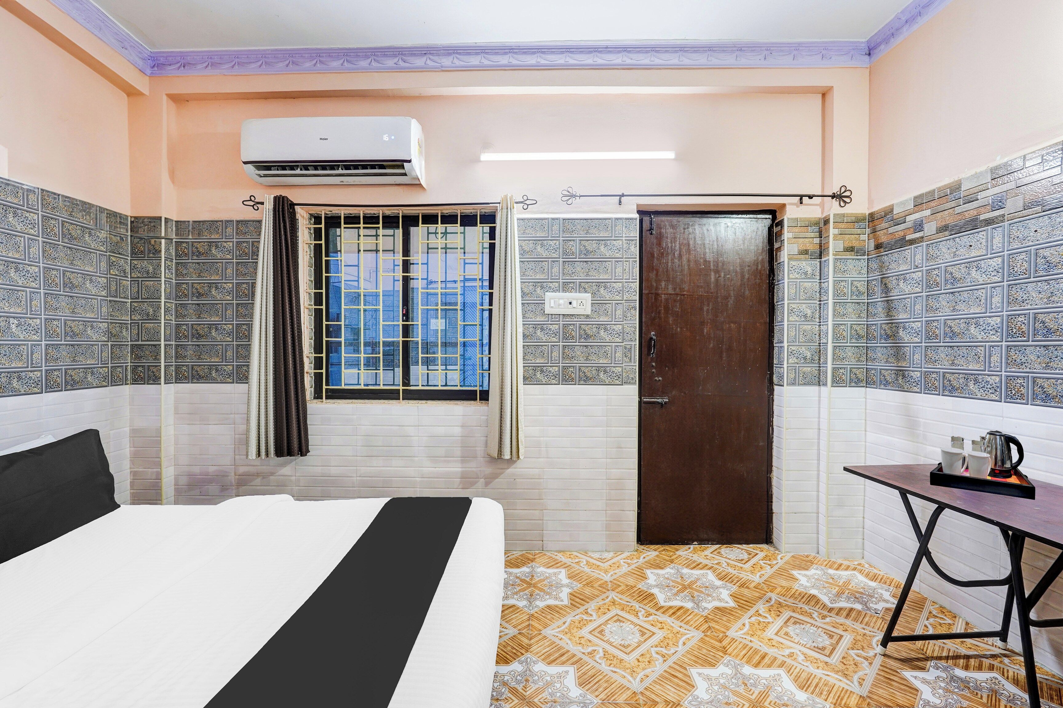 A hotel room with a bed, a window with curtains, a door, and a small table with a kettle and cups.
