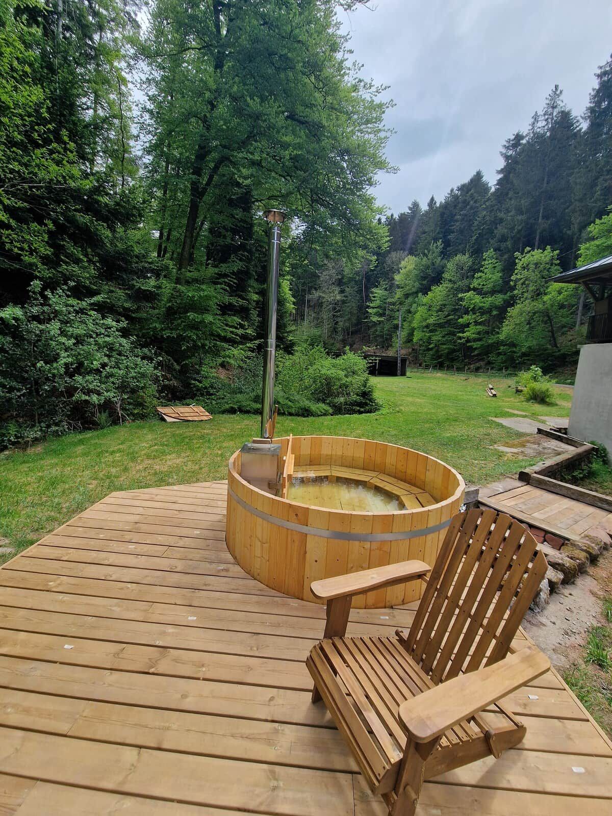 Un patio in legno con una vasca idromassaggio, una sedia di legno e uno sfondo boschivo.
