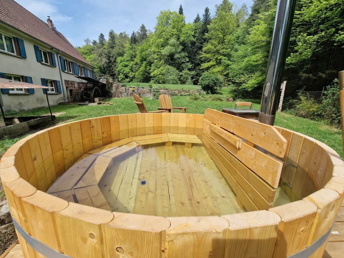Una vasca idromassaggio in legno con posti a sedere, circondata da un molo di legno, in un giardino sul retro con una casa e alberi sullo sfondo.
