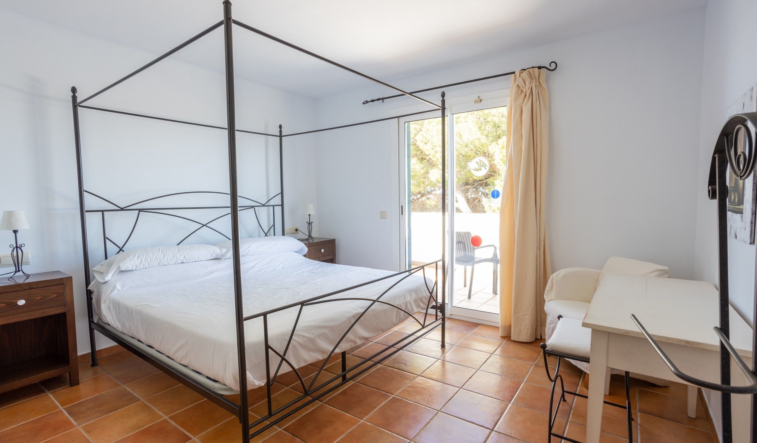 Una cama con dosel con una mesita de noche, un escritorio y una silla en una habitación con piso de baldosas y vistas por un vidrio.