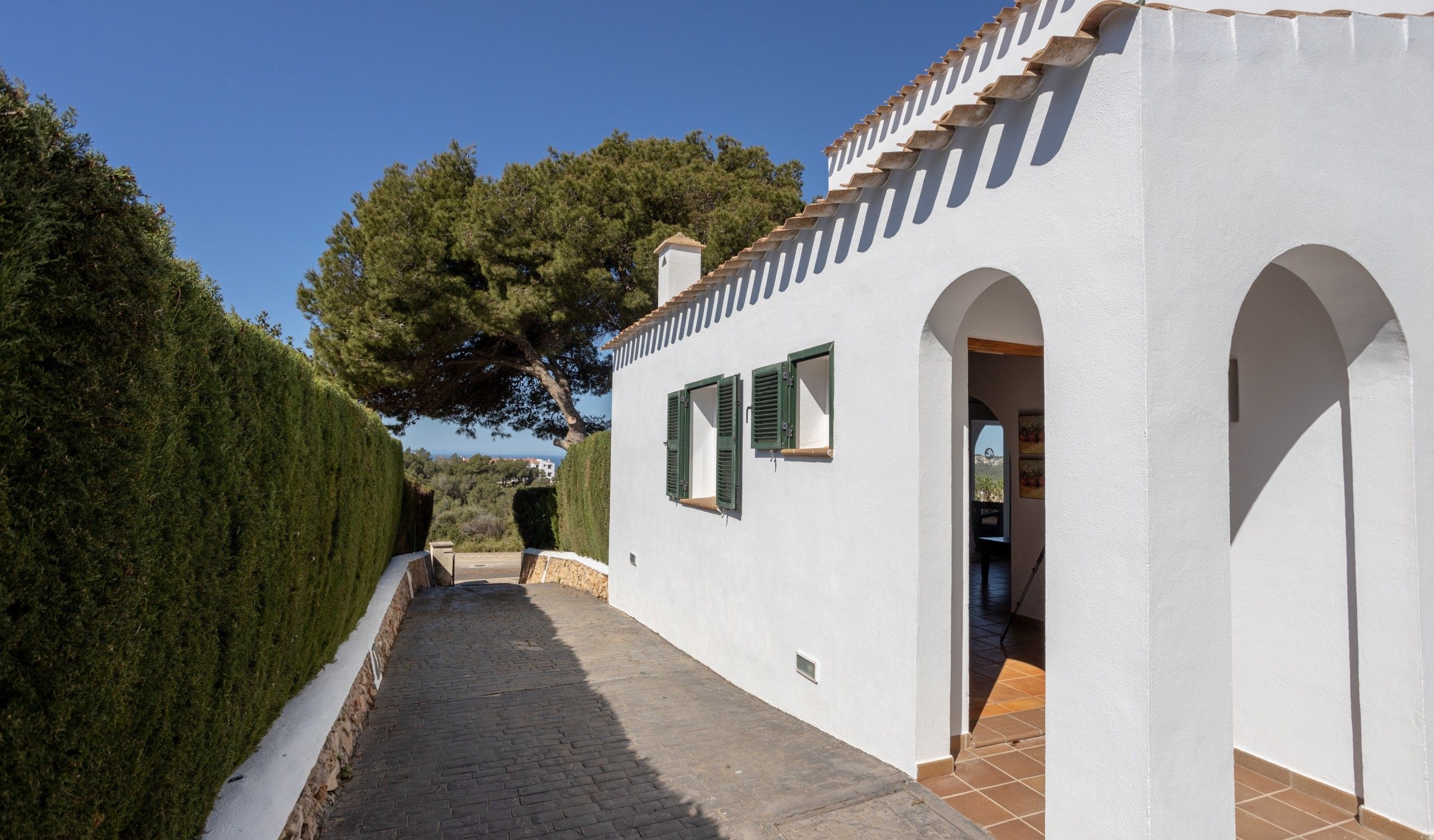A pathway leading to a white building with green shutters and a tiled roof.
