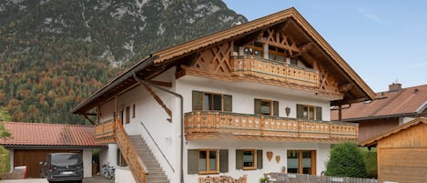 A two-story chalet with a wooden balcony, a staircase, and a mountain backdrop.