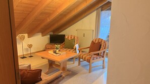 Ein gemütliches Dachzimmer mit Holzbalkendecke, einem Fernseher, einem Tisch mit Pflanze und zwei gemusterten Stühlen.