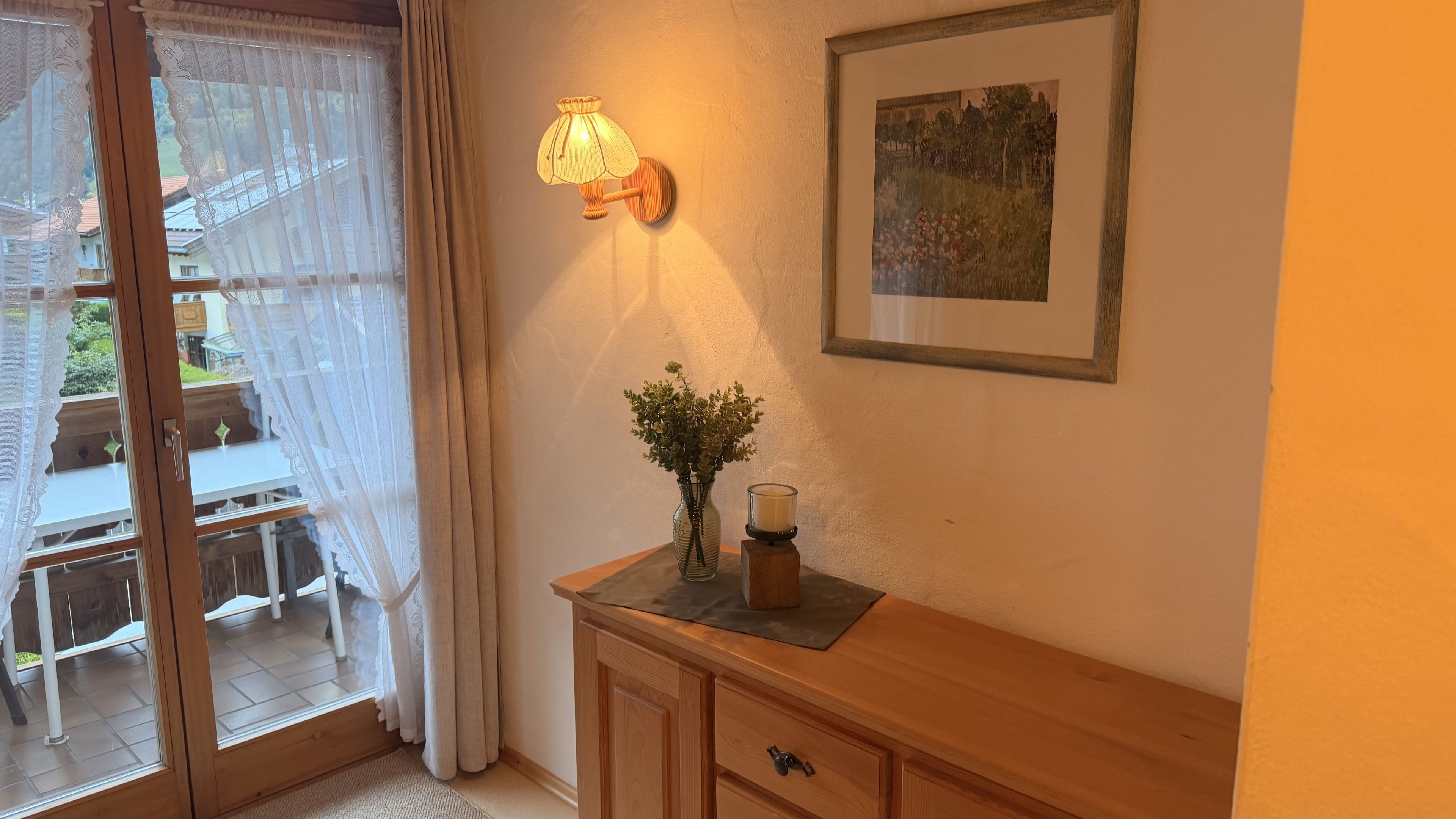 Ein Zimmer mit einem Holzkabinett, einer Blumenvase, einer Kerze, einer Wandlampe und einem gerahmten Bild.