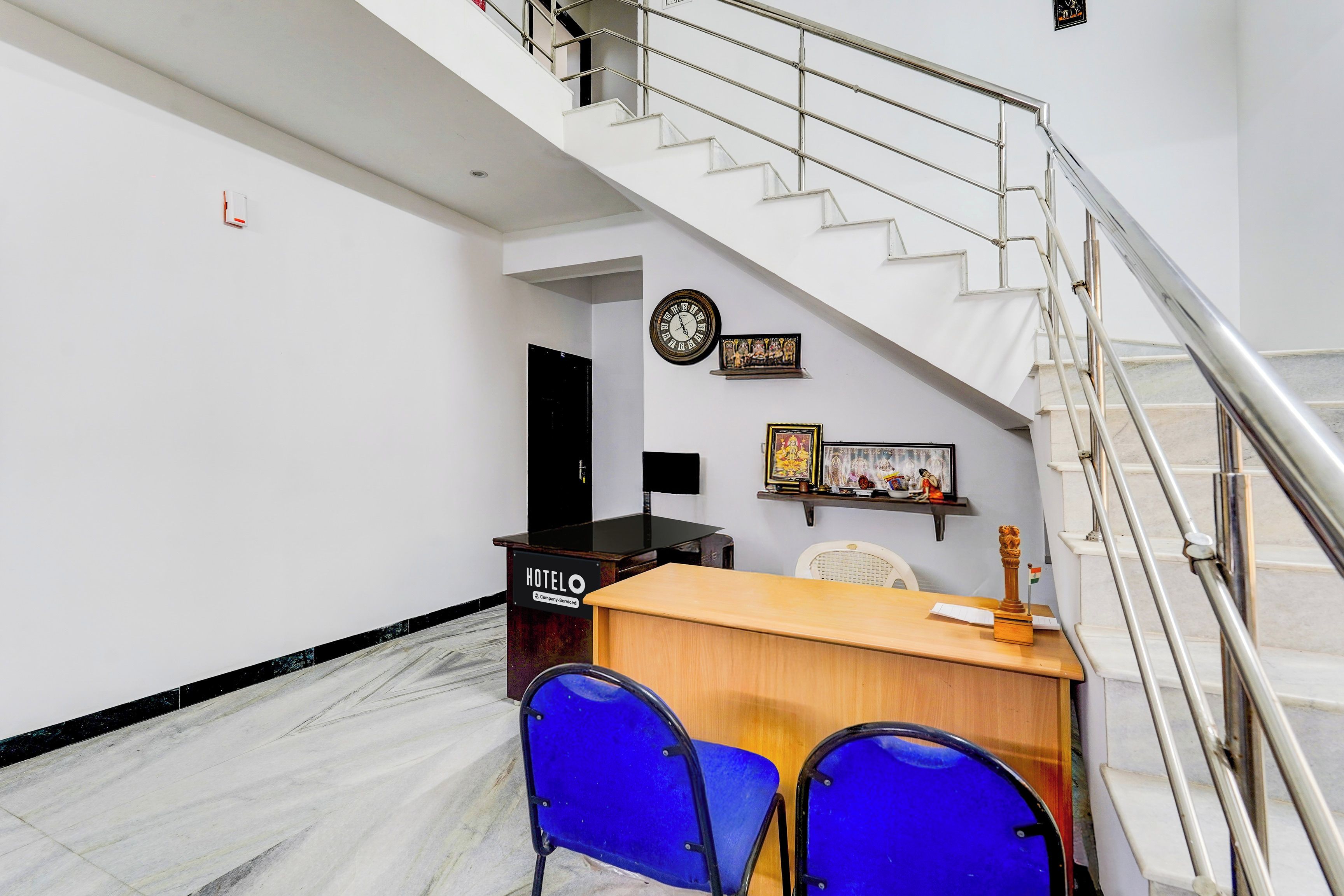 Zona de recepción de hotel con escritorio, dos sillas azules, una escalera y un reloj en la pared.