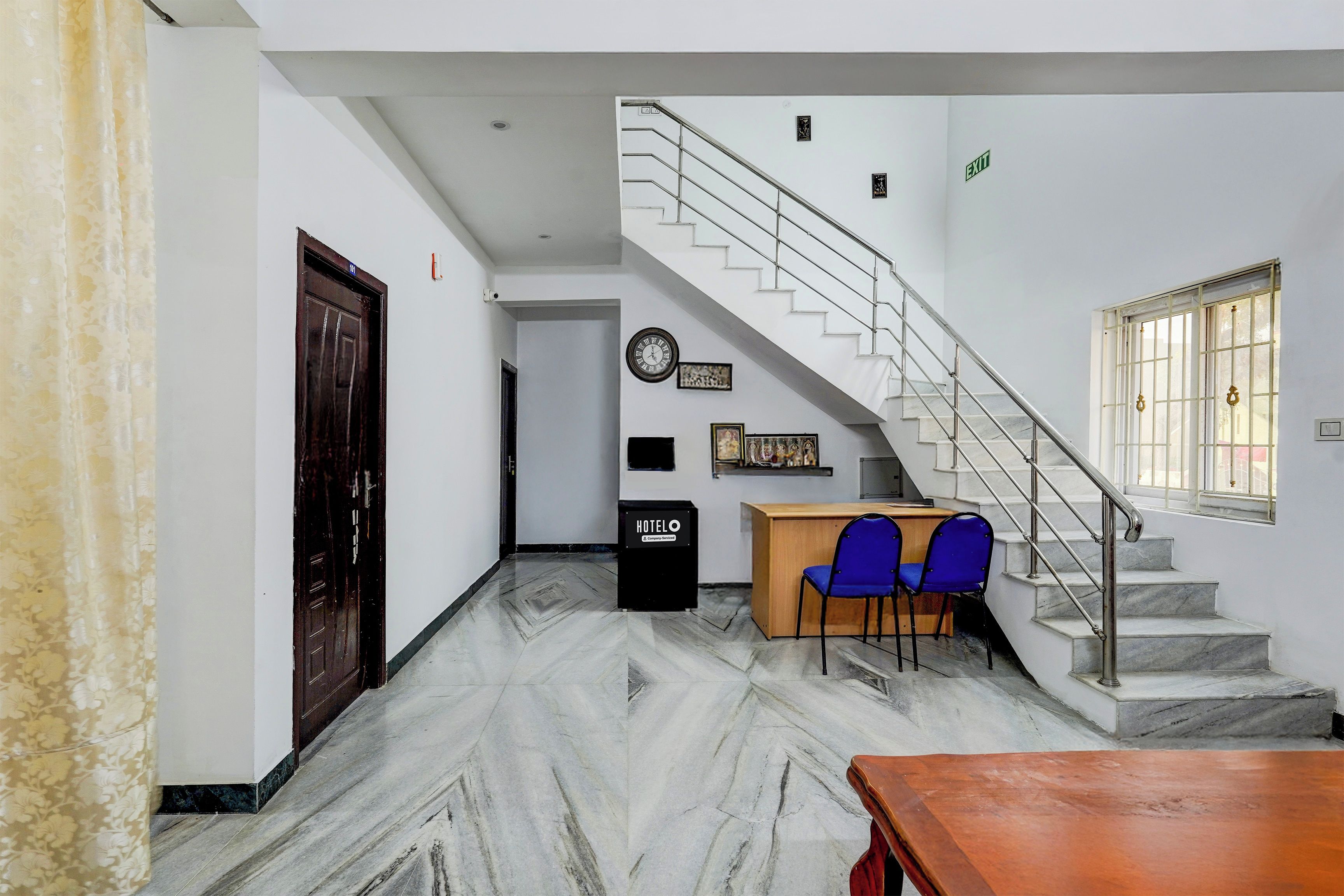 Vestíbulo de hotel con suelo de mármol, un mostrador de recepción, dos sillas azules, una mesa de madera, una escalera y un reloj en la pared.