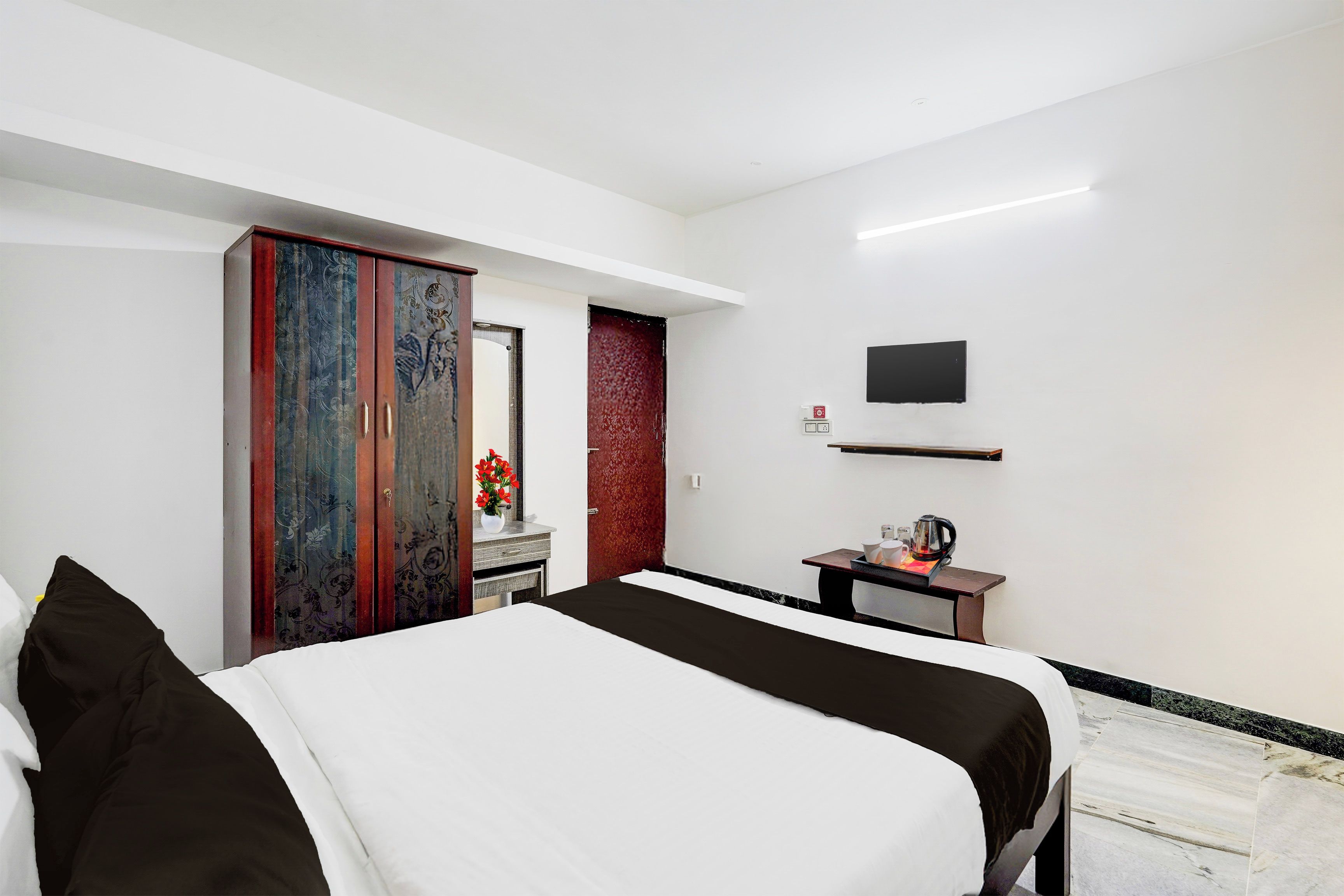 A hotel room with a bed, a wardrobe, a desk with a kettle, a flat-screen TV, and a red accent wall.