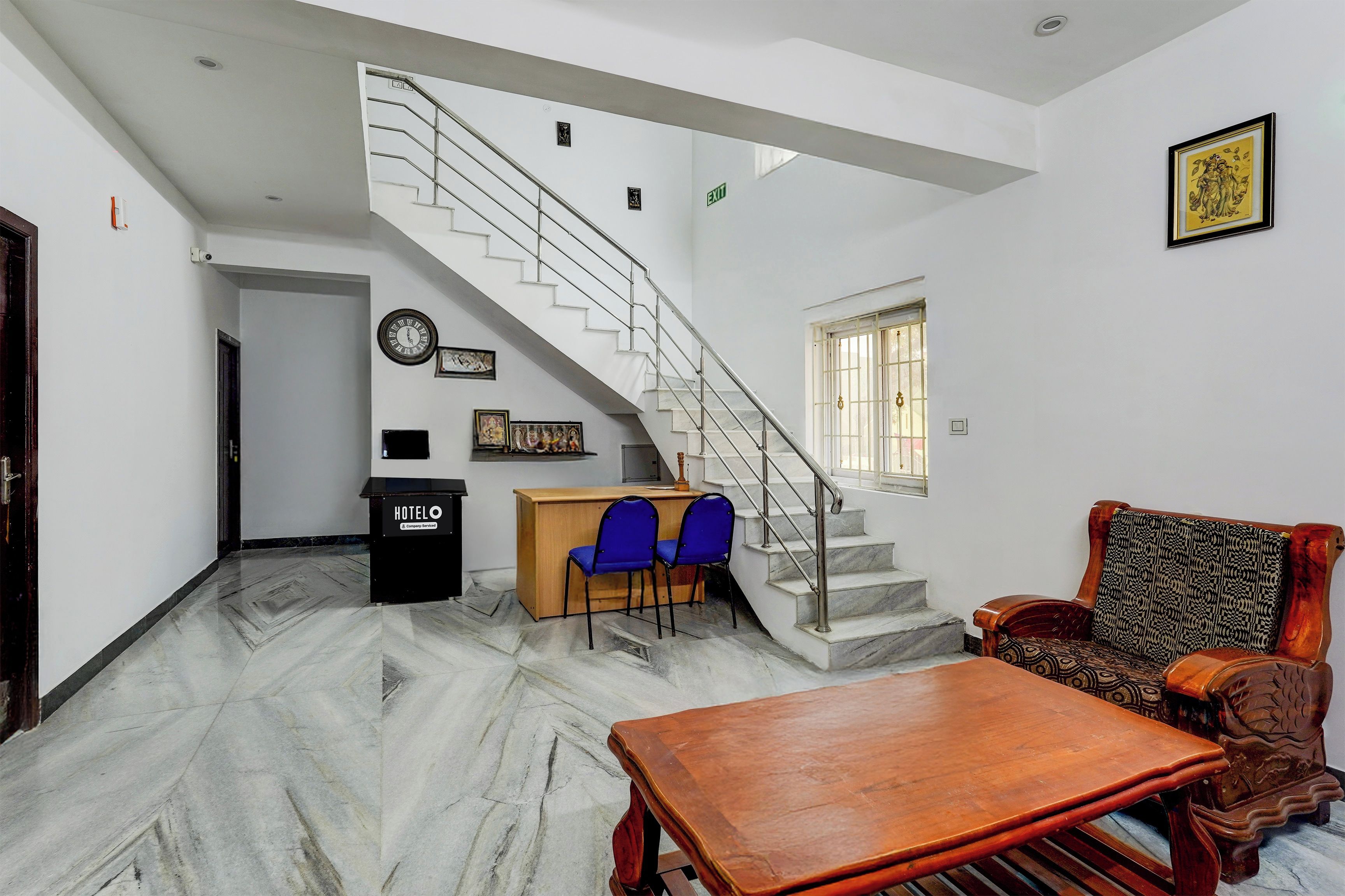 A hotel lobby with a marble floor, a wooden reception desk, blue chairs, a staircase, and a patterned sofa.