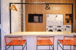 Un espace dédié à la lessive, propre et sec, avec un planning affiché, trois machines à laver et deux chaises orange.