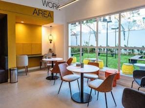A modern indoor dining area with round tables and chairs, a yellow accent wall, and large windows offering a view of the outdoors. - Nex One Sena Madureira (São Paulo)