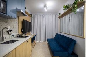 Une cuisine compacte dotée d’un téléviseur intégré, un coin repas avec un canapé bleu et une grande fenêtre avec des rideaux.