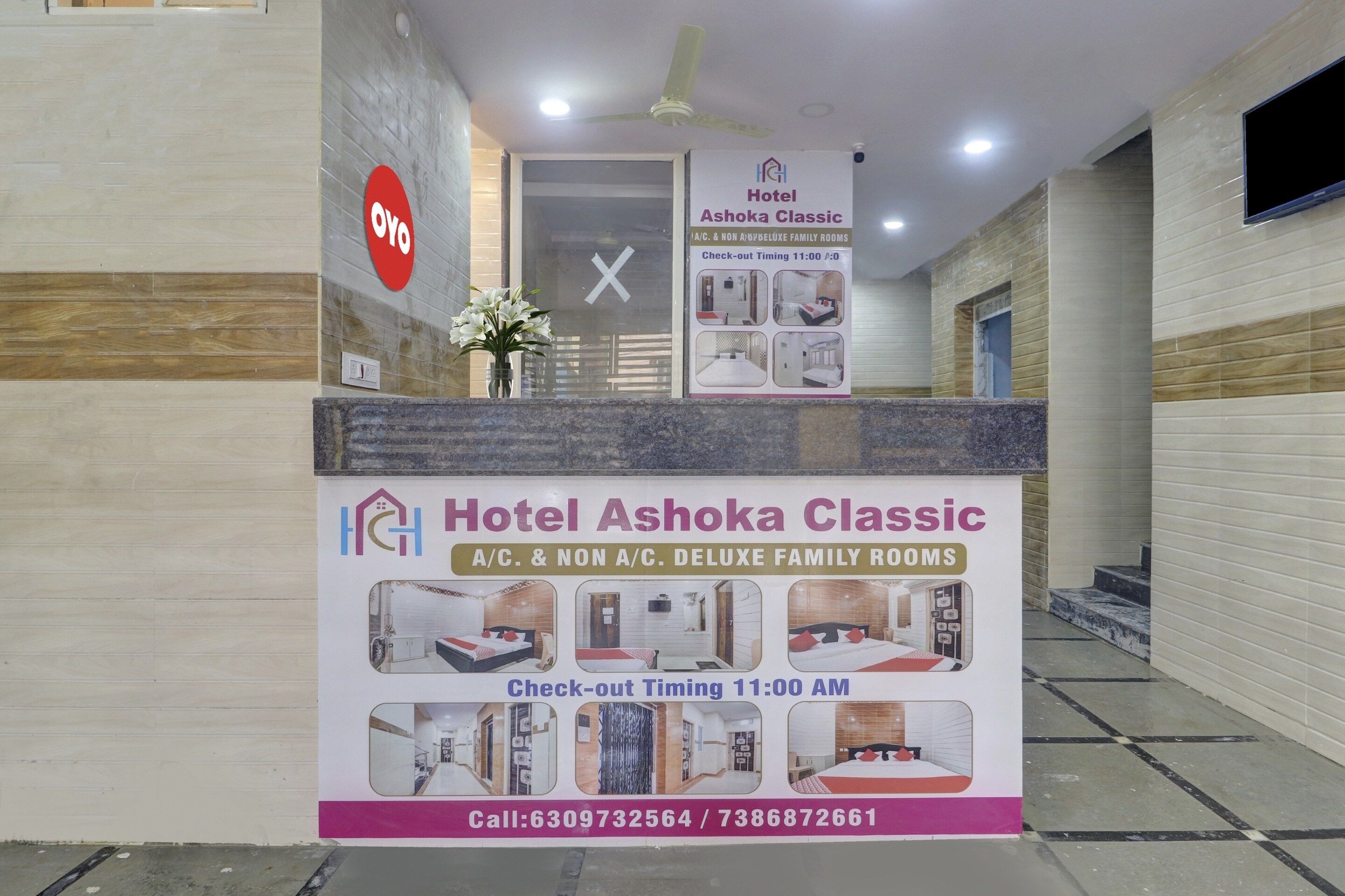A hotel reception desk with a sign for Hotel Ashoka Classic, indicating A/C. & non A/C. deluxe family rooms, check-out timing at 11:00 AM, and contact numbers.