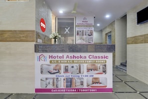 Una recepción de hotel con un cartel que anuncia Hotel Ashoka Classic, indicando habitaciones familiares de lujo con aire acondicionado y sin aire acondicionado, horario de salida a las 11:00 AM y números de contacto.