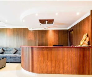 A reception area with a curved wooden reception desk, a ceiling fan, and a sofa in the background. - Deluxe Vaishnavi Residency (Hyderabad)