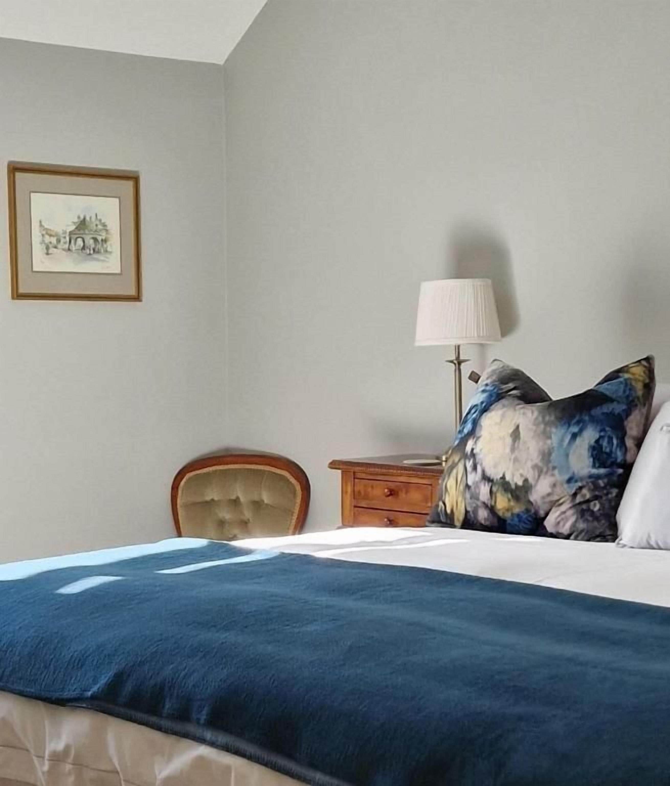 A bedroom with a bed, a chair, a nightstand, a lamp, and a framed picture on the wall.