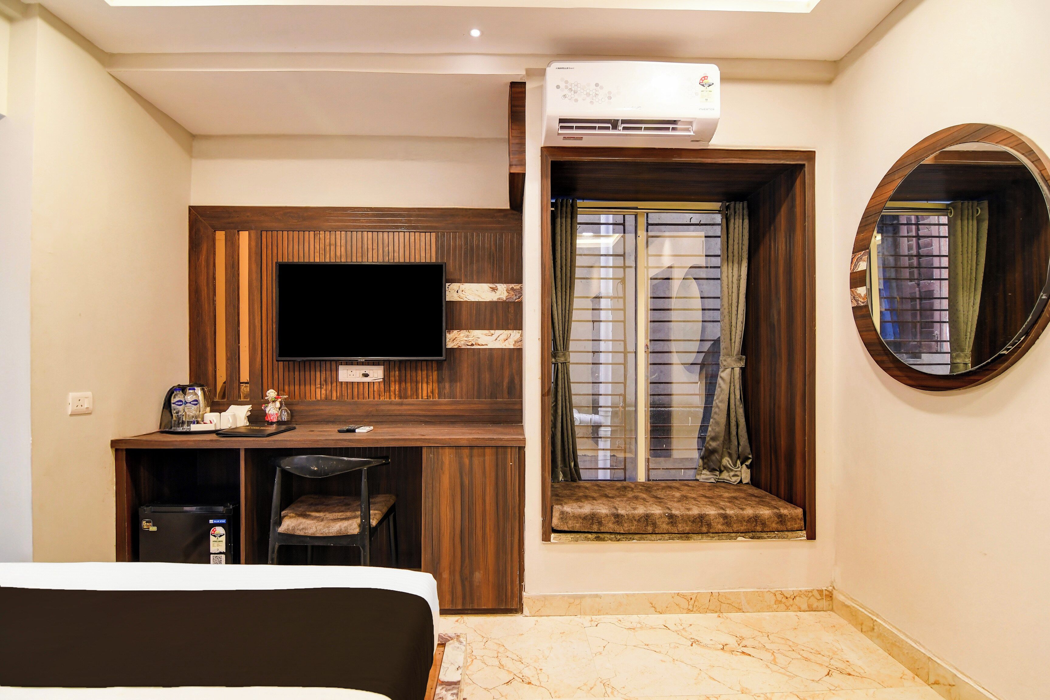 A hotel room with a bed, a desk with a flat-screen TV, an air conditioning unit, a window with a view, and a round mirror.