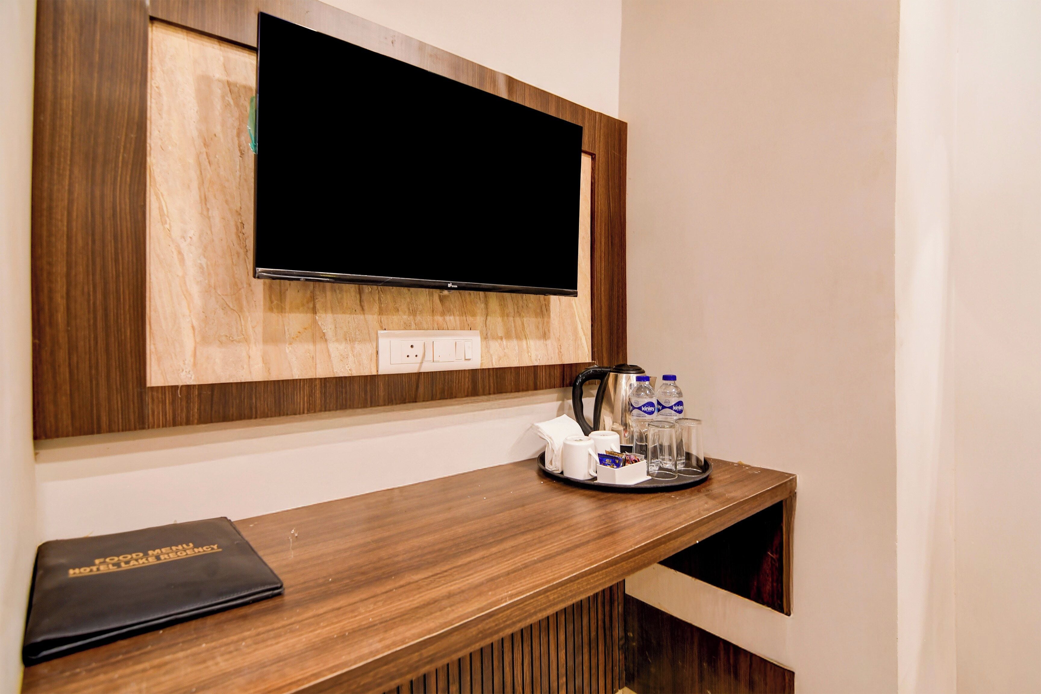 A hotel room with a mounted flat-screen TV, a wooden console table with a tray holding a kettle, glasses, and bottled water, and a black booklet labeled 'Food Menu Hotel Lake.'