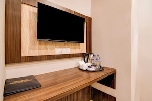 Habitación de hotel con televisor de pantalla plana montado, una mesita de madera con una bandeja que contiene una tetera, vasos y agua embotellada, y un folleto negro con la etiqueta 'Menú de Comidas Hotel Lake'.