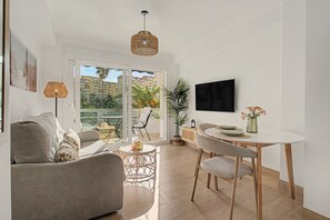 A modern living room with a sofa, a dining table, chairs, a TV, and a balcony with a view of buildings and palm trees.
