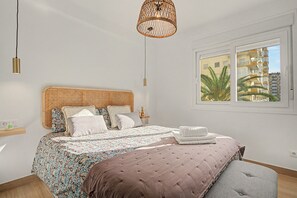A bedroom with a bed, a bedside table, a hanging light fixture, and a window with a view of buildings.