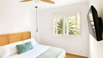 Un dormitorio con cama, ventilador de techo, televisión y ventana con vistas a palmeras.