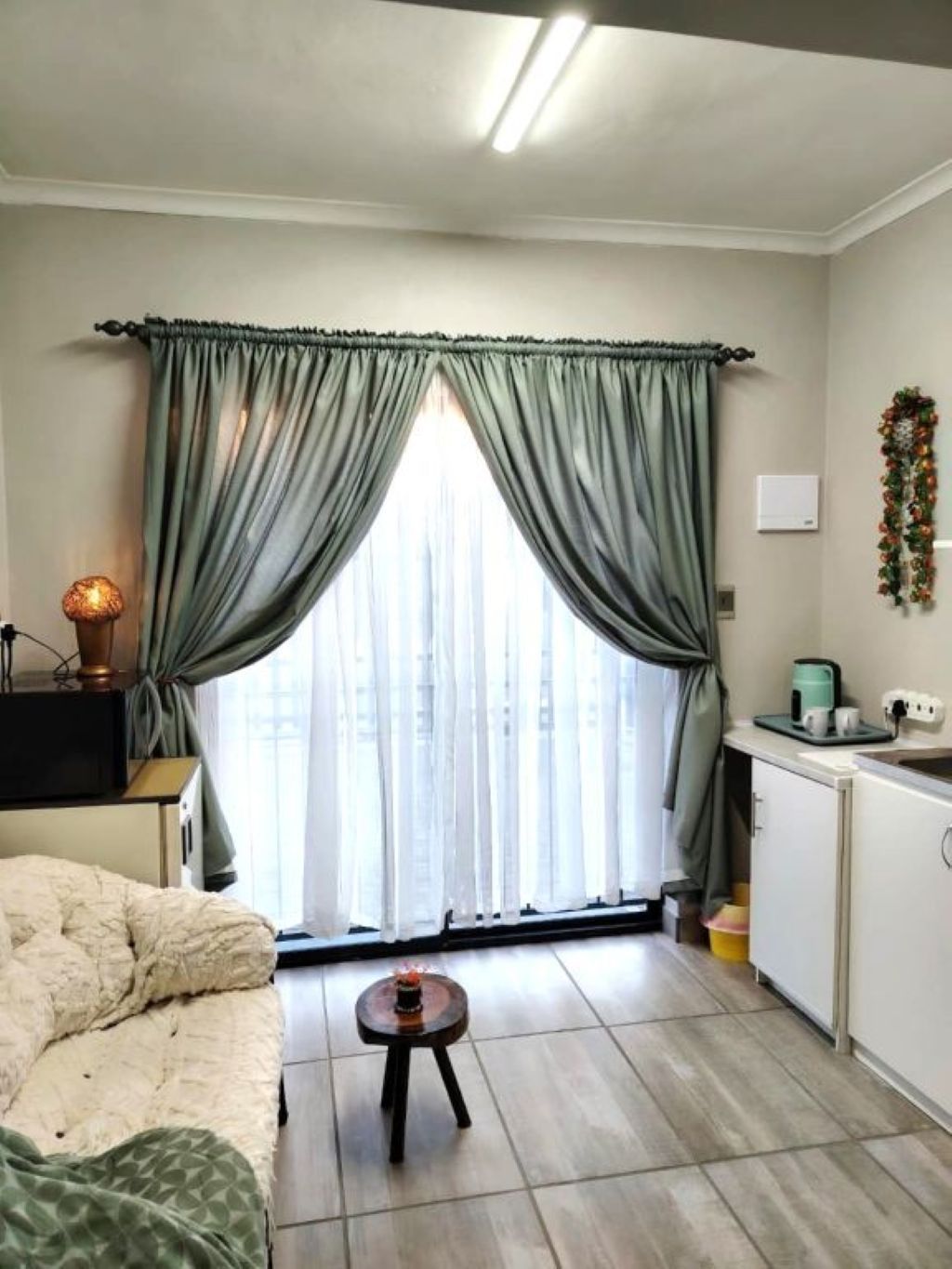 Una habitación compacta con cama, una mesita, un ventanal con cortinas, una kitchenette y una lámpara fijada a la pared.