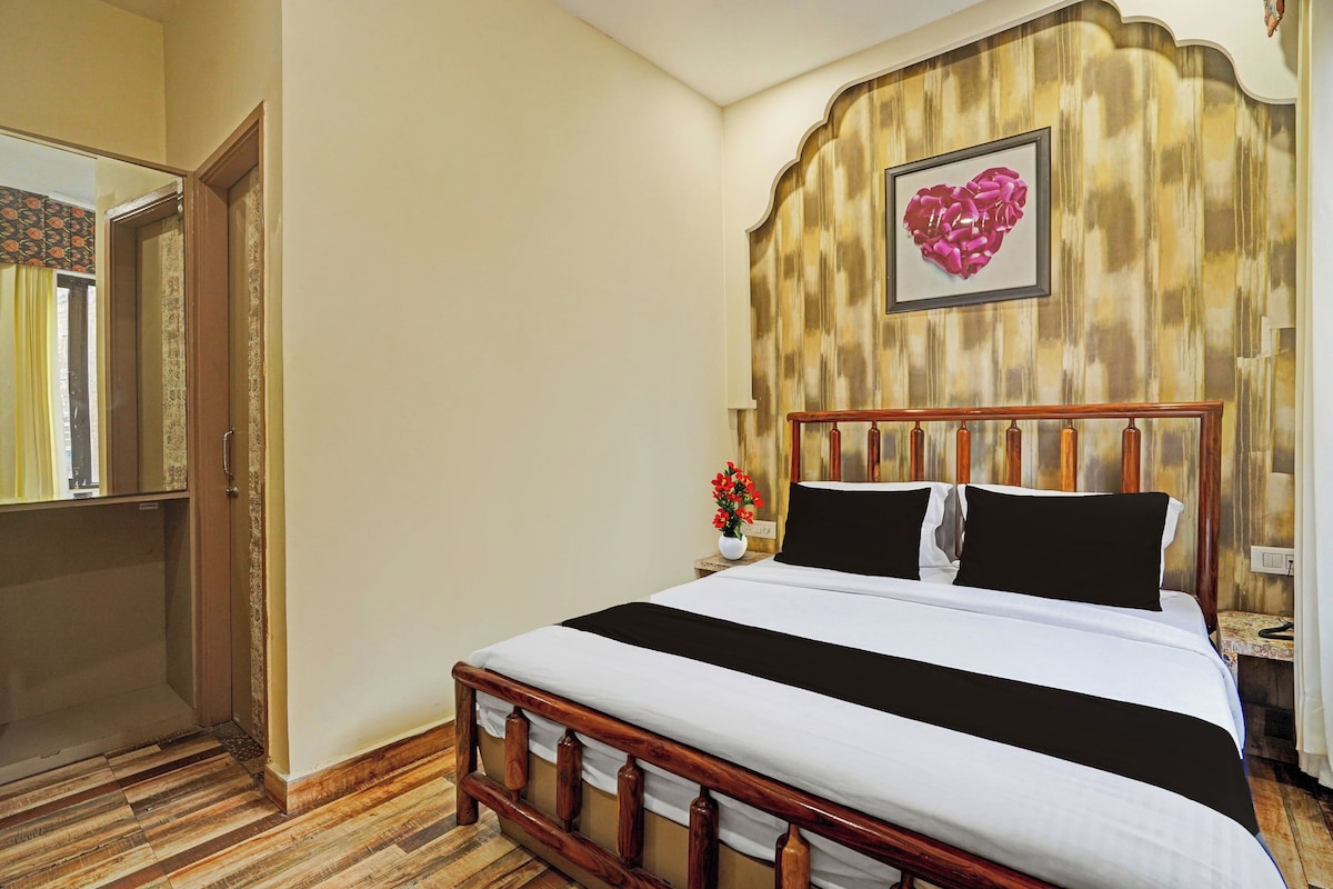 Quarto de hotel com cama de madeira, uma obra de arte em formato de coração na parede e uma mesa pequena com um vaso de flores.